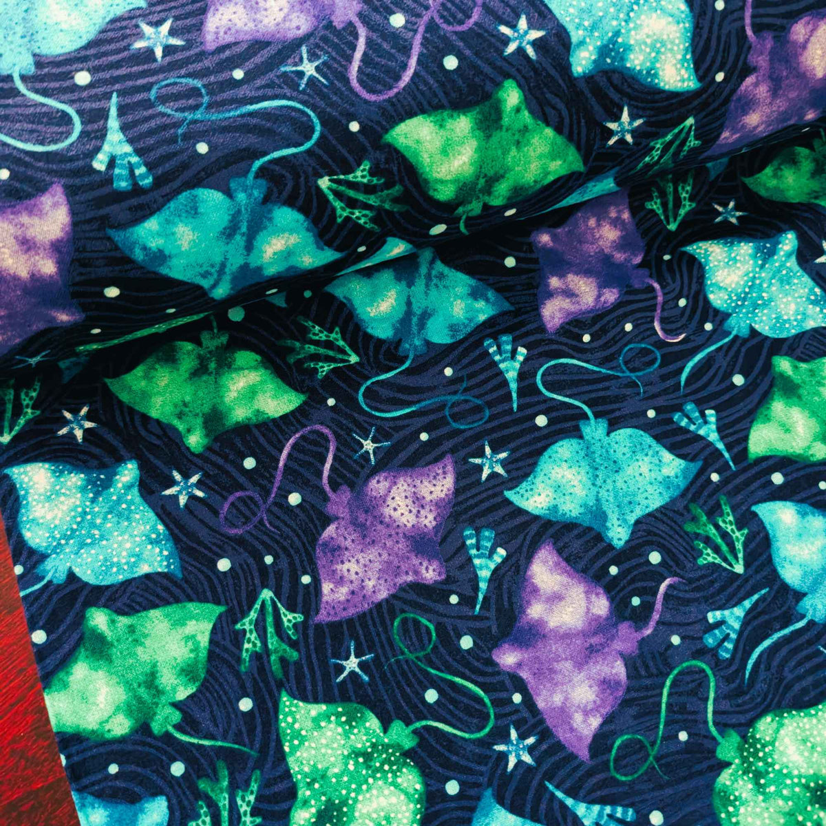 Cotton flannel fabric with colorful stingrays in blue, green, and purple on a dark ocean background with seaweed and starfish. Brushed soft, OEKO-TEX certified, 44 inches wide.