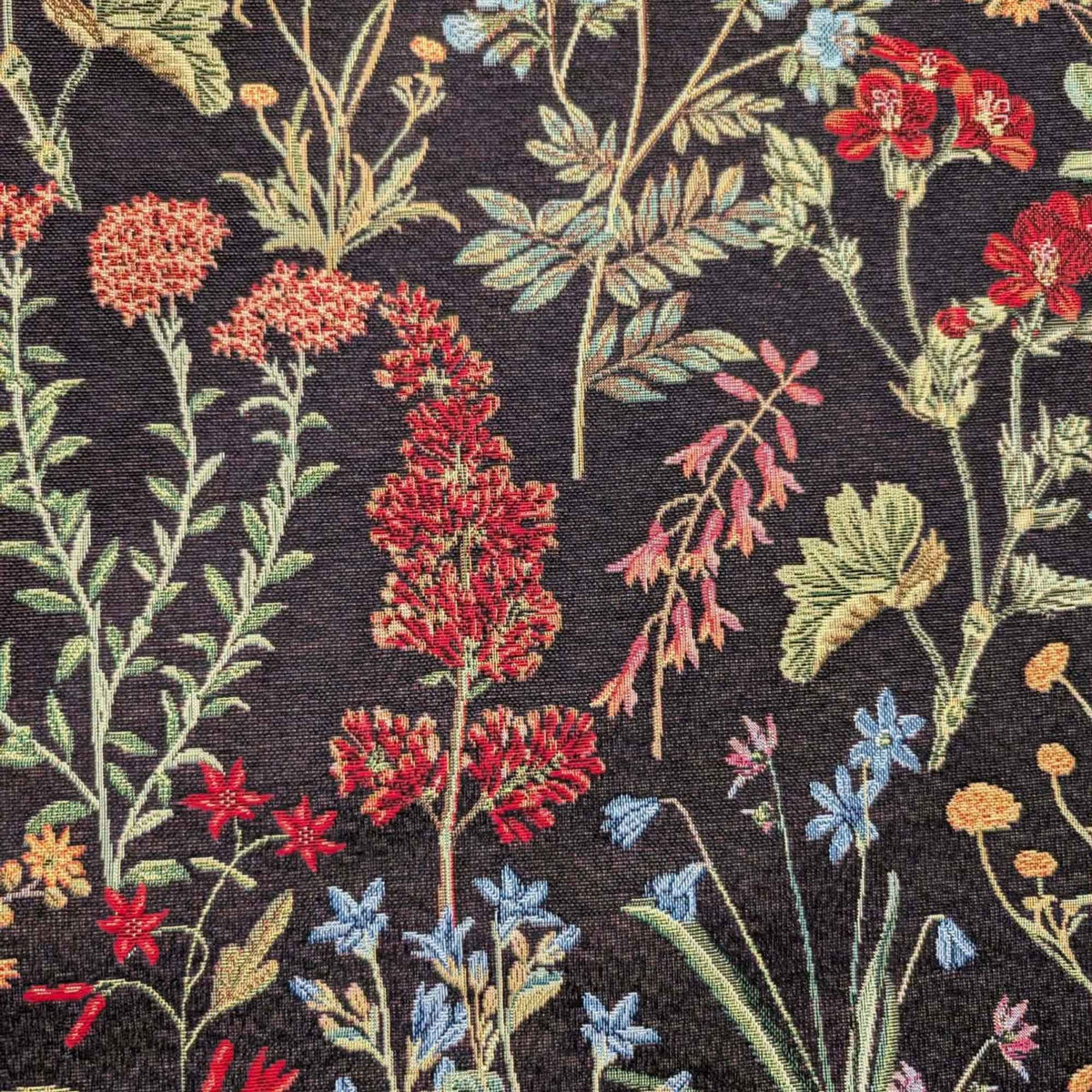 Botanical wildflower Gobelin tapestry fabric with red, coral, blue and yellow flowers and sage green leafy stems on a black background.