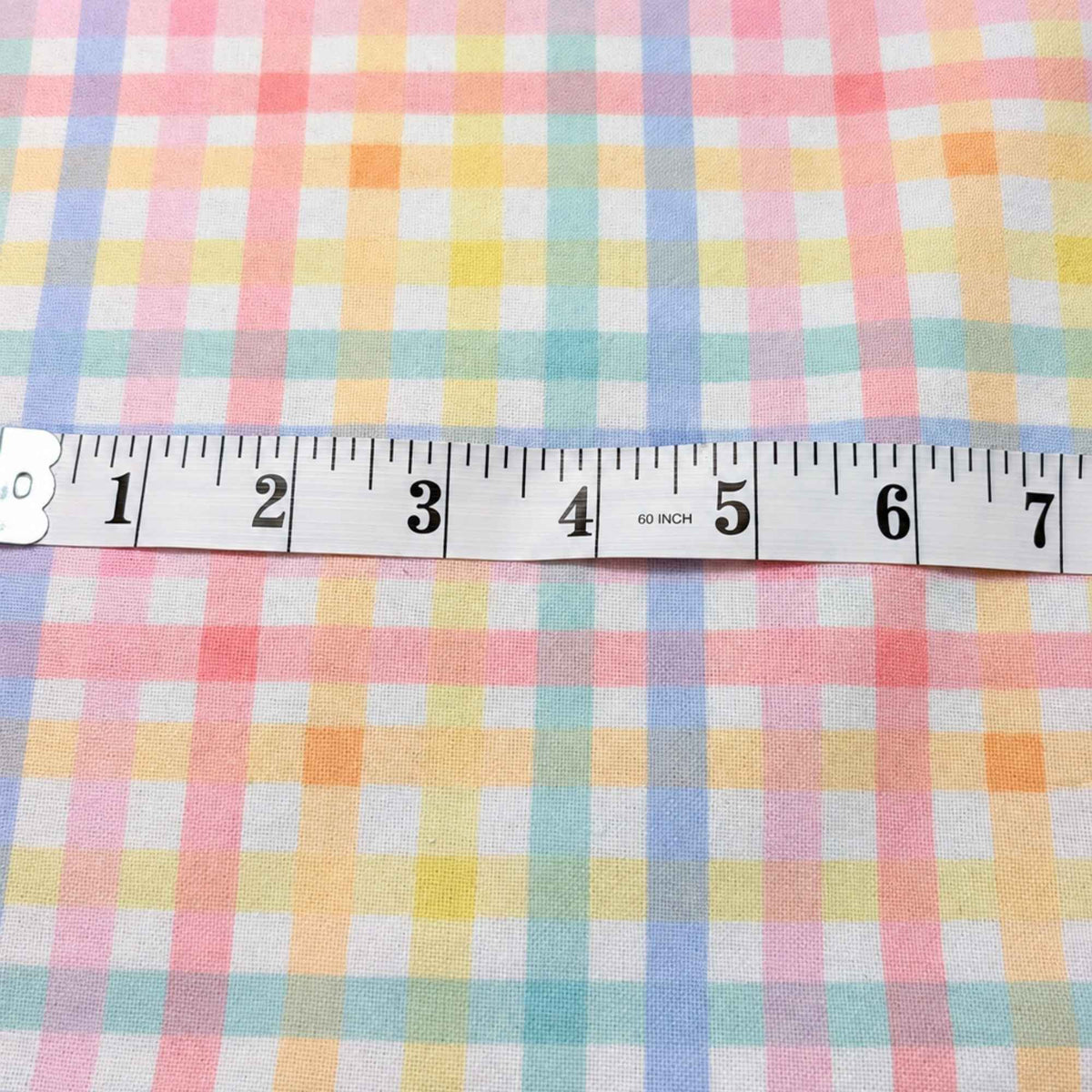 A close-up of pastel plaid fabric with a white measuring tape laid horizontally across the center to show the pattern scale. The fabric features a soft multicolor check pattern with pink, yellow, blue, green, and orange on a white background.