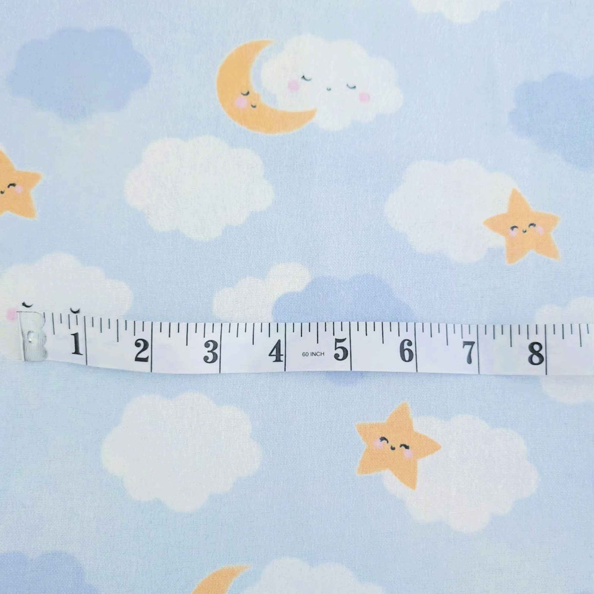 Light blue fabric with a playful pattern of smiling clouds, stars, and a crescent moon, shown with a white measuring tape to indicate scale.