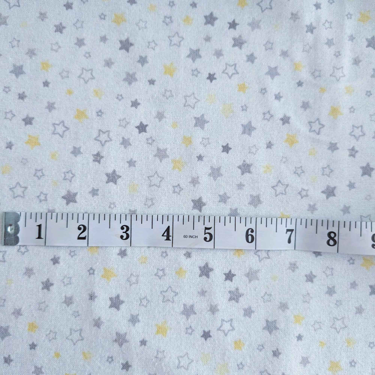 White fabric with a repeating pattern of small gray and yellow stars, shown with a metal tape measure across the middle for scale.