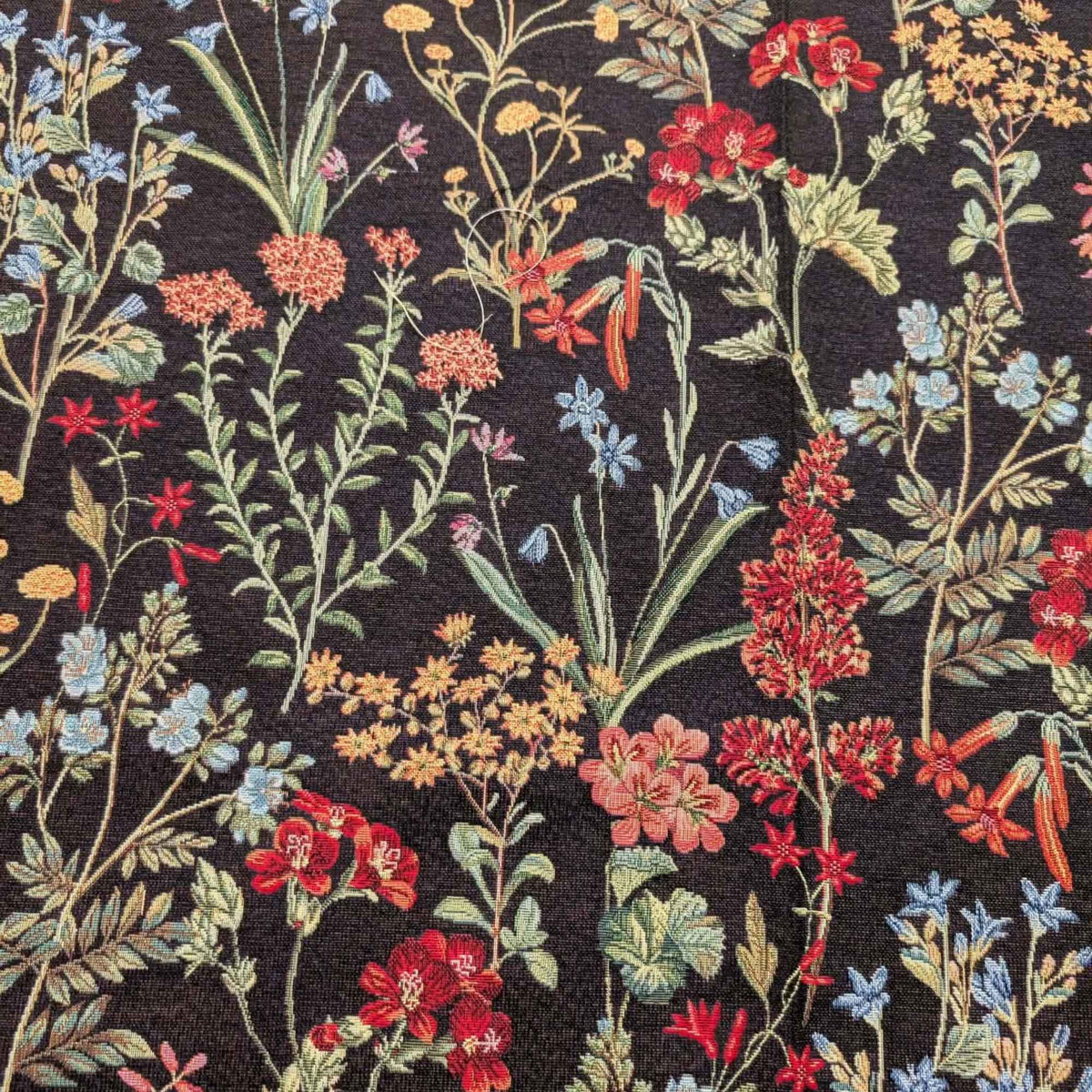 Botanical wildflower Gobelin tapestry fabric with red, coral, blue and yellow flowers and sage green leafy stems on a black background.